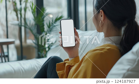 woman cozy living room holds smartphone, showcasing blank screen. serene atmosphere is enhanced by indoor plants and natural woman cozy living room holds smartphone, showcasing blank screen. serene atmosphere is enhanced by indoor plants and natural 124187927