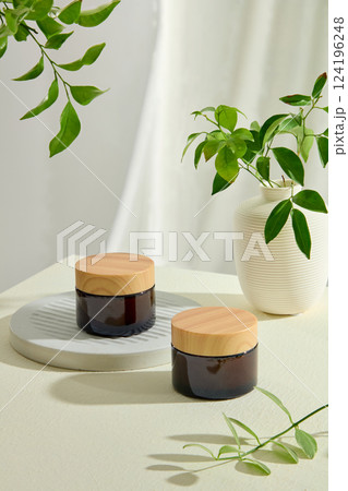 Brown cosmetic jar with yellow lid placed on stone pedestal, other jar placed on white surface. Decorative branches and leaves around. Bulb vase with green branches. Empty space for text. Brown cosmetic jar with yellow lid placed on stone pedestal, other jar placed on white surface. Decorative branches and leaves around. Bulb vase with green branches. Empty space for text. 124196248