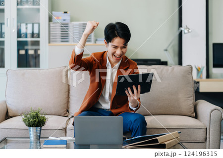 Happy young businessman celebrating success, raising hands in excitement while working on a laptop. 124196315