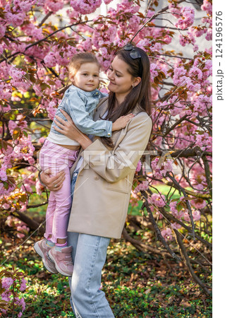 Young mother and daughter are standing in the park near a sakura tree that has blossomed with pink flowers in the spring in Prague. Young mother and daughter are standing in the park near a sakura tree that has blossomed with pink flowers in the spring in Prague. 124196576