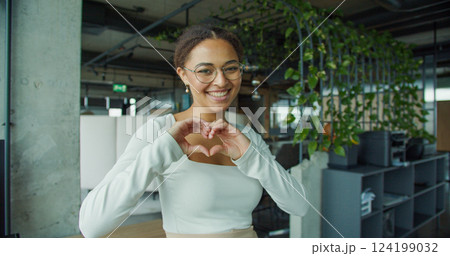 Cheerful young woman with glasses and curly hair smiling joyfully while making a heart shape with her hands, standing in a modern indoor office space with green plants and bright lighting 124199032