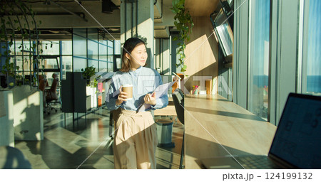 A young professional woman holding a coffee cup and clipboard in a modern office environment with large windows and natural light, emphasizing productivity and focus 124199132