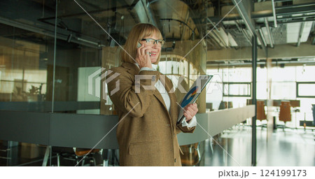 A professional businesswoman in a modern office environment, confidently discussing on the phone while holding a clipboard with data charts, standing near a glass partition A professional businesswoman in a modern office environment, confidently discussing on the phone while holding a clipboard with data charts, standing near a glass partition 124199173