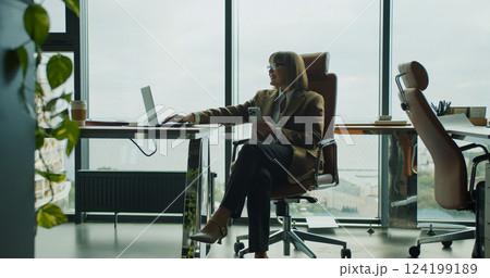 Relaxed businesswoman using smartphone in a modern office with large glass windows and a view of the city and sea in the background 124199189