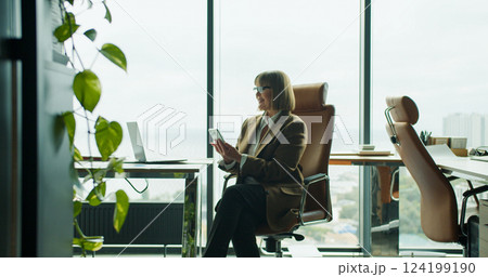 A focused businesswoman using smartphone while sitting in a modern office with a large window view overlooking the city and greenery A focused businesswoman using smartphone while sitting in a modern office with a large window view overlooking the city and greenery 124199190