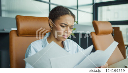 focused young woman analyzing documents at her desk in a modern office, surrounded by charts, a laptop, and office supplies with stylish leather chairs 124199259