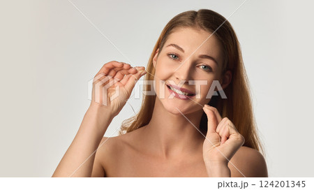 Smiling young woman with radiant skin using dental floss, emphasizing healthy teeth and oral hygiene in soft natural lighting 124201345