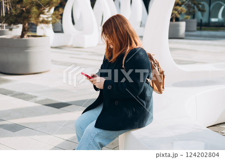 Happy woman chatting on phone and relaxing outdoors. Urban, people concept. Smiling 30s redhead woman text messaging on smart phone at sunny day on the city street, spring time 124202804