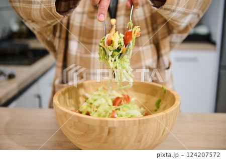 Person Tossing Fresh Salad Ingredients in Wooden Bowl in Kitchen Person Tossing Fresh Salad Ingredients in Wooden Bowl in Kitchen 124207572