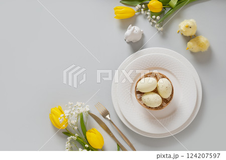 Easter table setting with spring yellow tulips, white plates, bunny, eggs on gray background. Happy Easter celebration. Copy space. View from above. Easter table setting with spring yellow tulips, white plates, bunny, eggs on gray background. Happy Easter celebration. Copy space. View from above. 124207597
