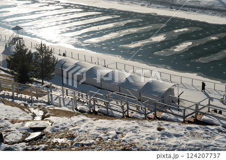 Winter landscape featuring a frozen river, snow-covered ground, and peaceful solitude near a quaint outdoor shelter in a serene and chilly environment 124207737