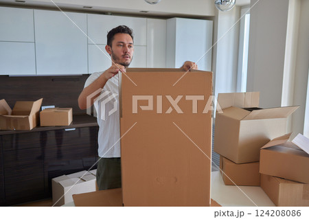 Man standing in living room on day of move to rented home, unbox carton box with belongings. Relocate to new own apartment collect personal things 124208086