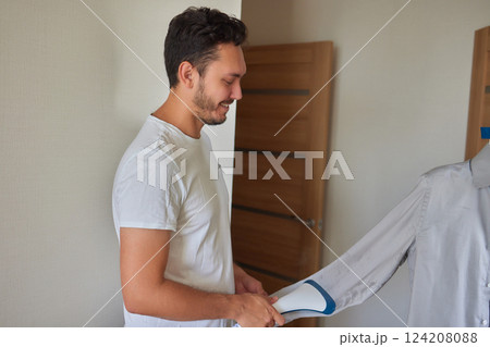 Domestic Device. Happy Middle Eastern Young Man Using Clothes Steamer And Steaming Shirt, Ironing His Clothes With Portable Gadget Domestic Device. Happy Middle Eastern Young Man Using Clothes Steamer And Steaming Shirt, Ironing His Clothes With Portable Gadget 124208088