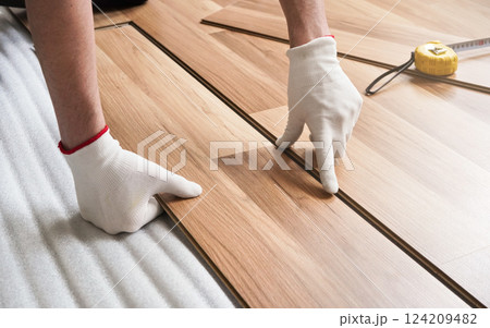 Installing laminated floor, detail on man hands in gloves fitting wooden tile, over white foam base layer, tape measure in background 124209482