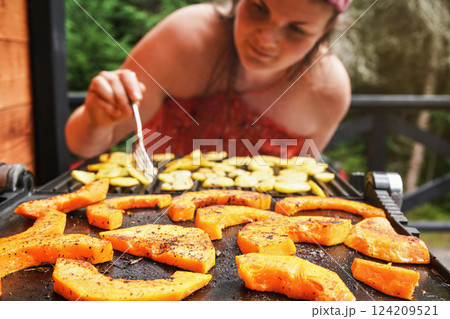 Butternut squash pieces grilled on electric grill, focus on bright orange vegetables seasoned with spice, blurred young woman in background 124209521