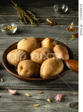 A wooden bowl contains several freshly harvested potatoes, surrounded by various spices and garlic on a textured rustic table, creating a warm culinary atmosphere A wooden bowl contains several freshly harvested potatoes, surrounded by various spices and garlic on a textured rustic table, creating a warm culinary atmosphere 124209814