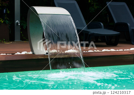Swimming pool with artificial waterfall in backyard of a luxury hotel on the tropics. Summertime vacation. Wooden floor near pool with blue water. 124210317