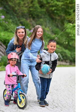 A happy group of people, including two women, a boy holding a ball, and a little girl on a balance bike with a helmet, enjoying outdoor activities on a paved path surrounded by greenery. A happy group of people, including two women, a boy holding a ball, and a little girl on a balance bike with a helmet, enjoying outdoor activities on a paved path surrounded by greenery. 124212583