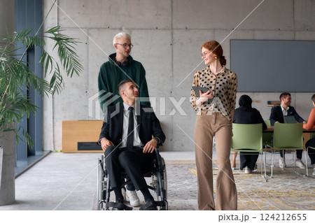 A diverse team of business colleagues, including a director in a wheelchair, engages in a collaborative discussion as they move together through a modern office space 124212655