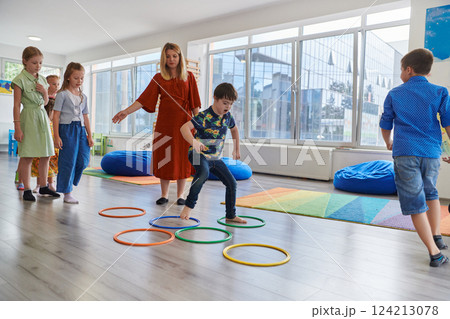 Small nursery school children with female teacher on floor indoors in classroom, doing exercise. Jumping over hula hoop circles track on the floor. Small nursery school children with female teacher on floor indoors in classroom, doing exercise. Jumping over hula hoop circles track on the floor. 124213078