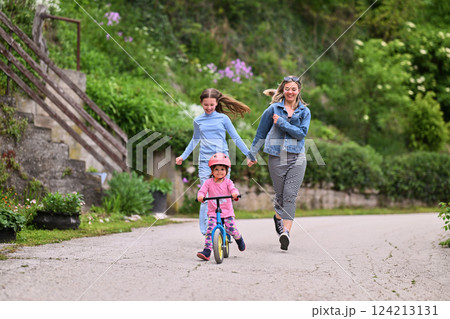 A cheerful little girl riding a balance bike with a helmet while two women joyfully run beside her on a paved path in a lush outdoor setting. 124213131