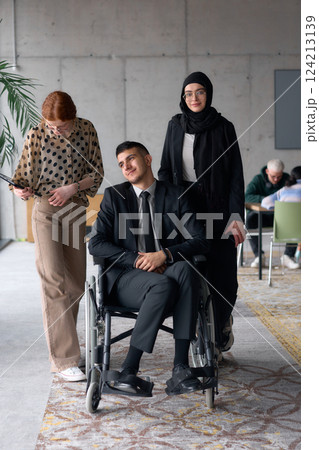 A diverse business team poses confidently, including a smiling director seated in a wheelchair, flanked by two professional female colleagues, one wearing a hijab and the other holding a tablet, in a 124213139