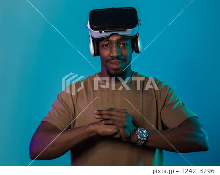 In an avant-garde scene, an African American man engages in cutting-edge virtual reality gaming, utilizing VR glasses to immerse himself in futuristic boxing games, set against a vivid blue background 124213296