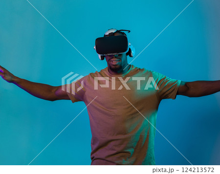 African American man dons VR glasses, engrossed in a virtual reality simulation, gesturing with hands, against a striking blue background, highlighting the fusion of cutting-edge technology and African American man dons VR glasses, engrossed in a virtual reality simulation, gesturing with hands, against a striking blue background, highlighting the fusion of cutting-edge technology and 124213572