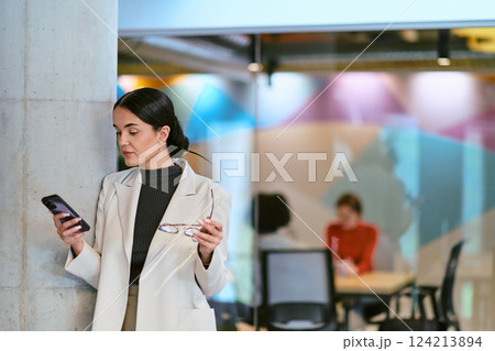 In a modern glass office, a contemporary businesswoman effortlessly multitasks, using her smartphone, while surrounded by colleagues, illustrating the dynamics of corporate life and technology 124213894