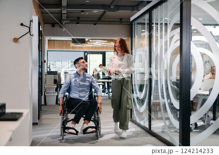 Business colleagues, collaborative business colleagues, including a person in a wheelchair, walk past a modern glass office corridor, illustrating diversity, teamwork and empowerment in the workplace. Business colleagues, collaborative business colleagues, including a person in a wheelchair, walk past a modern glass office corridor, illustrating diversity, teamwork and empowerment in the workplace. 124214233