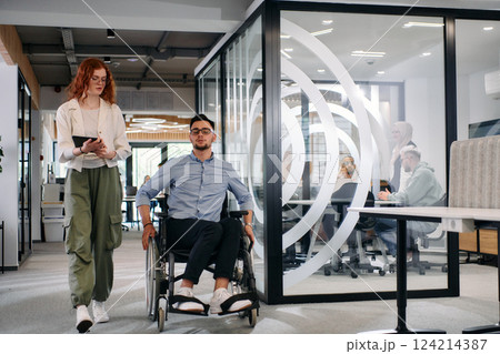 Business colleagues, collaborative business colleagues, including a person in a wheelchair, walk past a modern glass office corridor, illustrating diversity, teamwork and empowerment in the workplace. Business colleagues, collaborative business colleagues, including a person in a wheelchair, walk past a modern glass office corridor, illustrating diversity, teamwork and empowerment in the workplace. 124214387