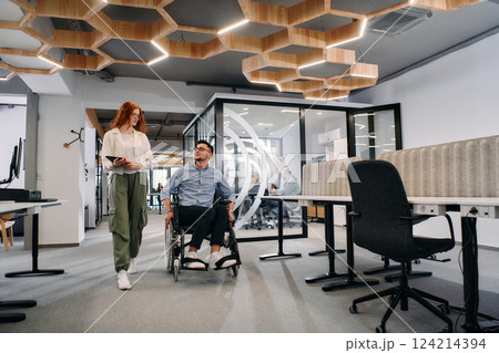 Business colleagues, collaborative business colleagues, including a person in a wheelchair, walk past a modern glass office corridor, illustrating diversity, teamwork and empowerment in the workplace. Business colleagues, collaborative business colleagues, including a person in a wheelchair, walk past a modern glass office corridor, illustrating diversity, teamwork and empowerment in the workplace. 124214394