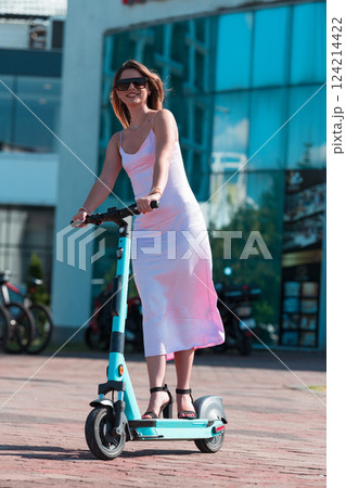Elegant woman in a pink dress and sunglasses riding an electric scooter through the sunny city streets, embracing the freedom and style of a perfect urban adventure. 124214422