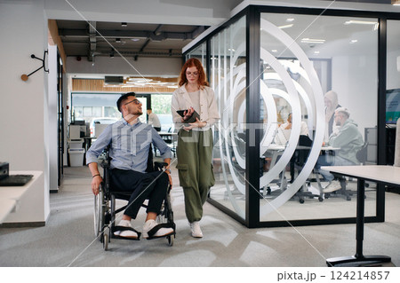 Business colleagues, collaborative business colleagues, including a person in a wheelchair, walk past a modern glass office corridor, illustrating diversity, teamwork and empowerment in the workplace. Business colleagues, collaborative business colleagues, including a person in a wheelchair, walk past a modern glass office corridor, illustrating diversity, teamwork and empowerment in the workplace. 124214857