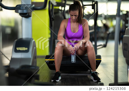 A fit woman takes a well deserved break in the gym, sipping water from her bottle after an intense workout session A fit woman takes a well deserved break in the gym, sipping water from her bottle after an intense workout session 124215308
