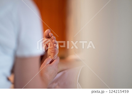 Foot Massage Therapy in a Relaxing Spa Setting. A professional massage therapist in a white uniform provides a soothing foot massage to a client in a calm and serene spa environment 124215459
