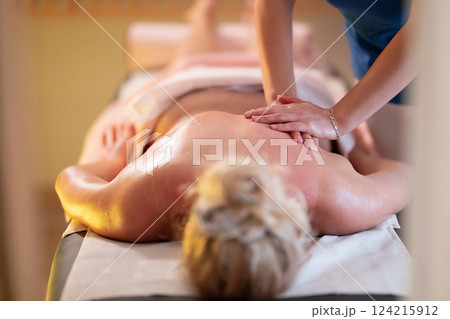 Relaxing Back Massage in a Spa Setting. A professional massage therapist in a blue uniform provides a relaxing back massage to a client lying on a massage table. 124215912
