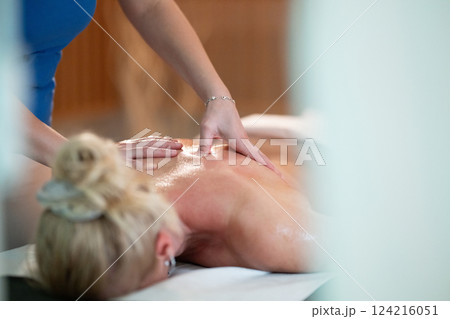 Relaxing Back Massage in a Spa Setting. A professional massage therapist in a blue uniform provides a relaxing back massage to a client lying on a massage table. 124216051