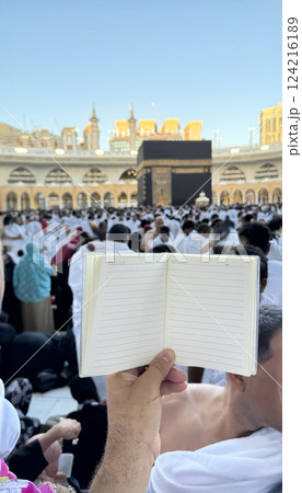 A pilgrim holds a small notebook with a heart doodle in front of the Kaaba in Mecca, Saudi Arabia. The sacred site is surrounded by worshippers performing Hajj or Umrah under a clear blue sky 124216189