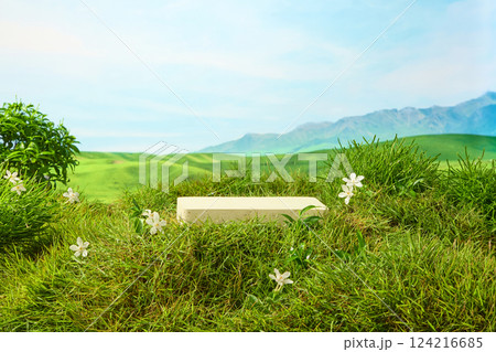 Rectangular stone pedestal with smooth flat surface placed on green grass. Tiny white petals and yellow pistil flowers grow lushly. Mountain scenery, blue sky. Empty space for showing product 124216685