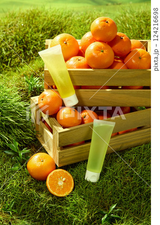 Tube of lotion sits on a wooden crate filled with vibrant oranges. The round Citrus stacked on top of each other. Oranges are cut in half to reveal a juicy surface with tiny seeds. Mockup for design  124216698