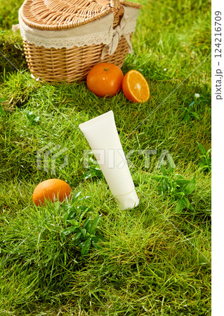 An unlabeled tube of cosmetics sits tilted on a green lawn with lush foliage. A ripe orange sits around it. The orange is cut in half and placed next to a bamboo picnic basket with a lace border. An unlabeled tube of cosmetics sits tilted on a green lawn with lush foliage. A ripe orange sits around it. The orange is cut in half and placed next to a bamboo picnic basket with a lace border. 124216709