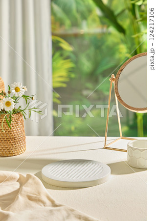 Empty stone pedestal portrait in the center of the photo. Round metal mirror on white table. Hand woven natural flower basket with little white flowers. Scene for advertising with minimal concept. Empty stone pedestal portrait in the center of the photo. Round metal mirror on white table. Hand woven natural flower basket with little white flowers. Scene for advertising with minimal concept. 124217106