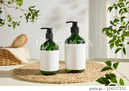 Two unlabeled green pump cosmetic bottles are placed on a round straw mat. A square straw basket contains a white towel and a wooden comb. Green leaves decorate the corners of the photo Two unlabeled green pump cosmetic bottles are placed on a round straw mat. A square straw basket contains a white towel and a wooden comb. Green leaves decorate the corners of the photo 124217132