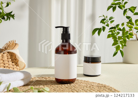 A towel is placed in a corner of the photo, a plastic pump bottle is displayed on a round straw mat. A brown plastic cosmetic jar has no label. Branches and leaves decorate the surrounding area A towel is placed in a corner of the photo, a plastic pump bottle is displayed on a round straw mat. A brown plastic cosmetic jar has no label. Branches and leaves decorate the surrounding area 124217133