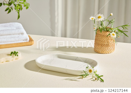 Oval white marble tray for cosmetics or jewelry. Blooming flowers in a handmade wicker vase. Wooden tray for neatly folded white towels, decorated by flowers around. Minimal scene for advertising. 124217144