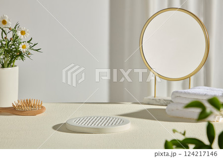 A white empty pedestal in the center to place products. A round gold mirror, white towels, a flower pot and a wooden comb are placed on the white table. Stand for displaying cosmetics or products. 124217146