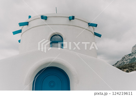 A blue door is on a white building. The building is small and has a blue roof. The sky is cloudy and the building is on a hill. 124219611