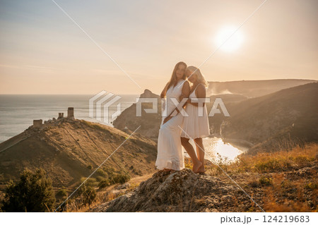 Two young girls are standing on a hillside, one of them wearing a white dress. The sun is shining brightly, creating a warm and inviting atmosphere. The girls seem to be enjoying their time together. 124219683