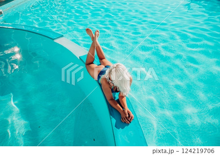A woman is laying on a pool ledge, wearing sunglasses and a blue bikini. She is smiling and she is enjoying her time at the pool. 124219706